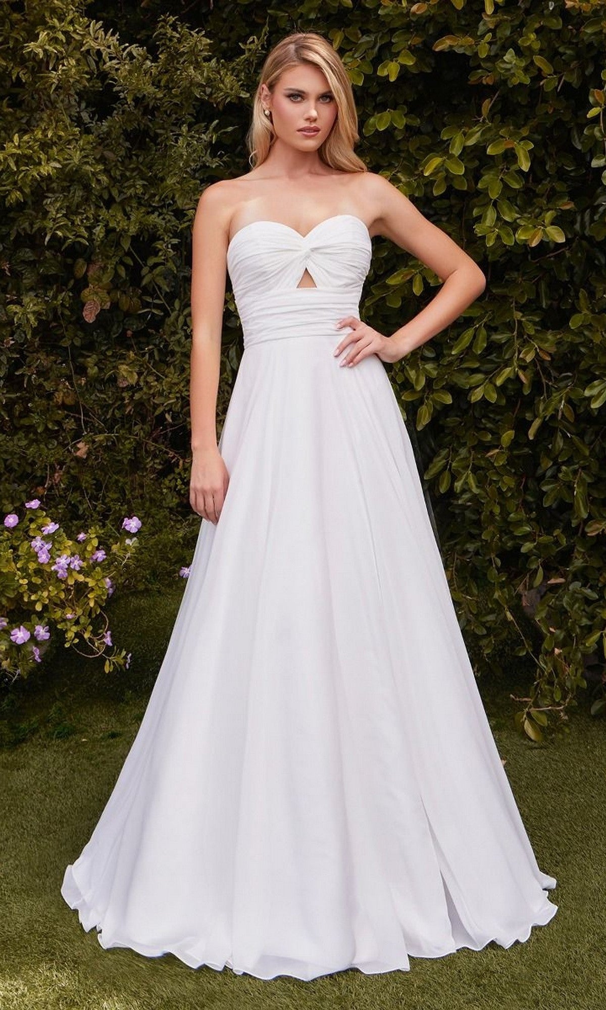 A woman poses outdoors against lush greenery in the Ladivine 7435W Strapless Long White Prom Gown, featuring a sweetheart neckline, front cutout, and off-white color—an affordable formal dress with one hand on her hip.